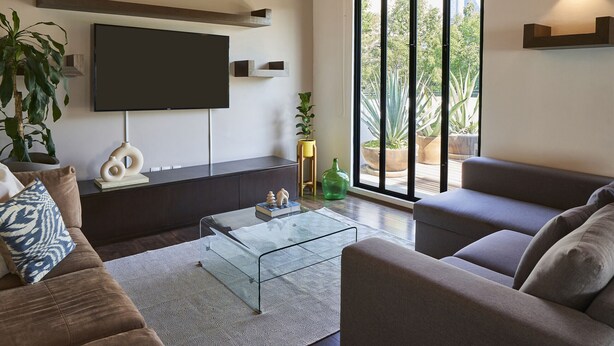 A modern living room with a flat-screen TV, comfortable seating, a coffee table, and large windows offering a view of the outdoors.
