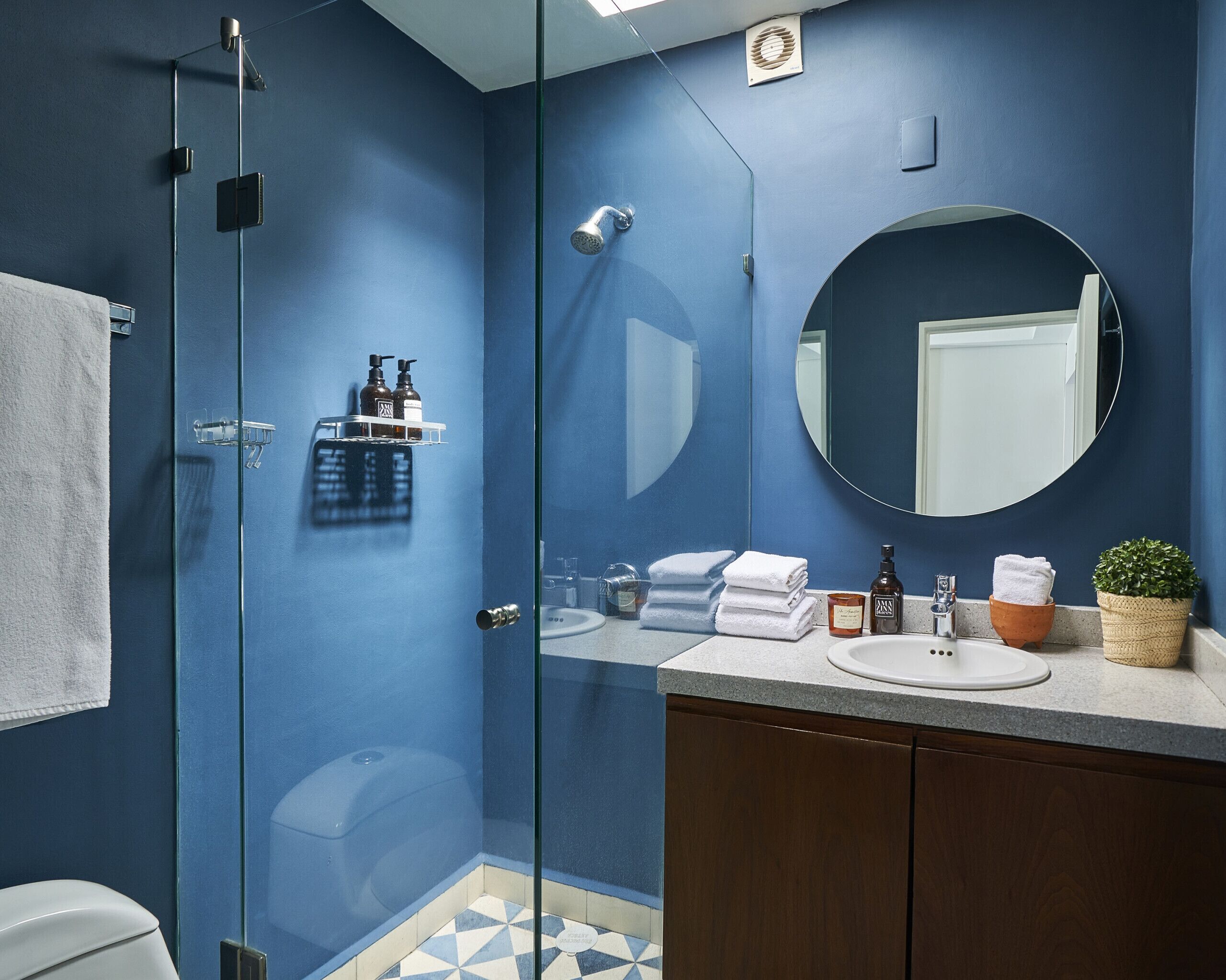 A modern bathroom with a blue wall, a glass shower enclosure, a round mirror, a sink with a countertop, and a wooden cabinet.