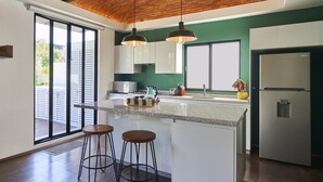 A modern kitchen with a large island, two bar stools, a refrigerator, and a view through the sliding glass doors.