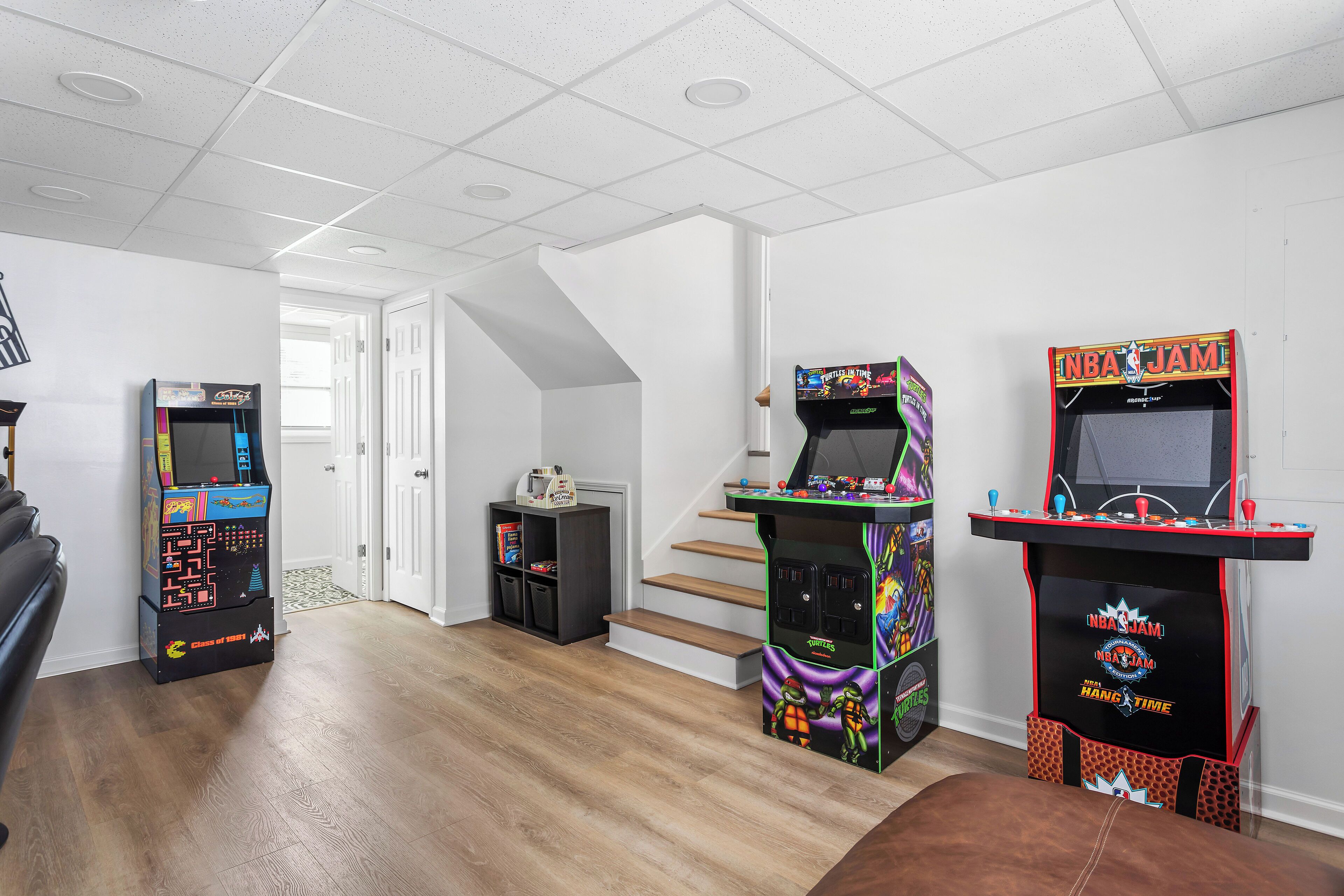 A room with wooden flooring, white walls, and a staircase. It contains two arcade machines, one labeled "NBA JAM" and another with a colorful design. There is a small shelf with books and a bag of chips.