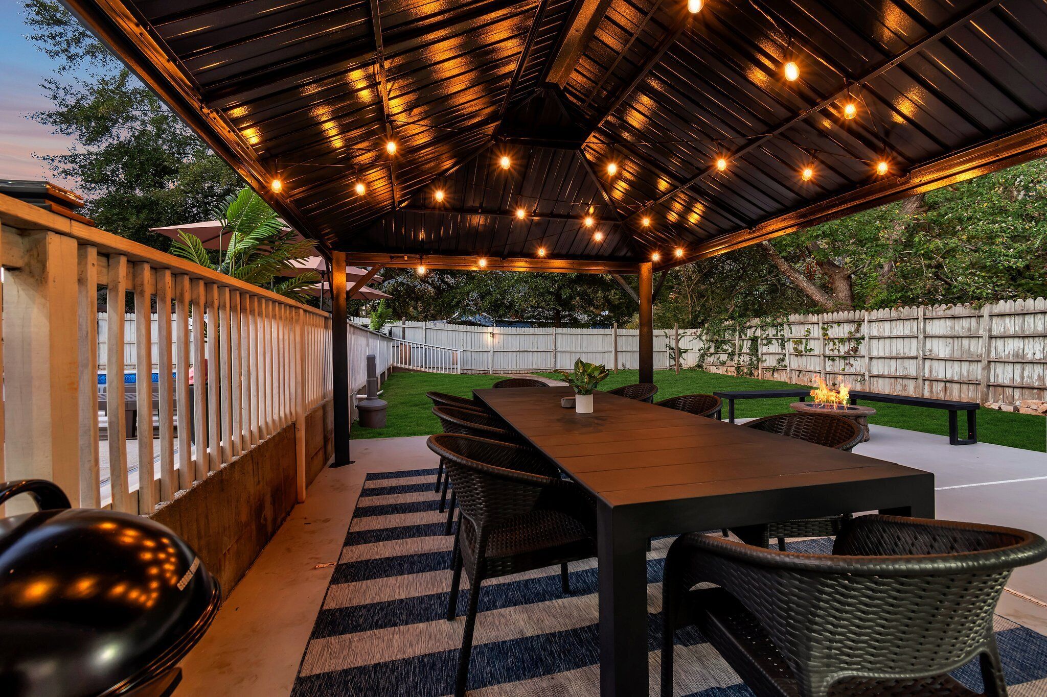 Eine Terrasse im Freien mit Holztisch und -stühlen, einem Grill und einem überdachten Bereich mit Lichterketten.