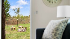A room with a view of a grassy outdoor area, a wooden deck, and a black couch with a patterned pillow. - Tiny Cottage - Hill Country (Kyle)