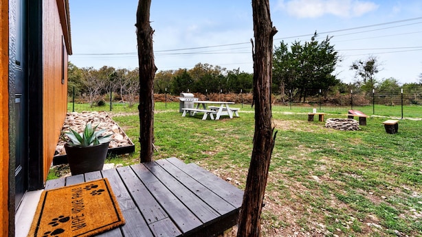 A wooden deck with a doormat, a picnic table, and a barbecue grill in a grassy outdoor area with trees in the background. - Tiny Cottage - Hill Country (Kyle)