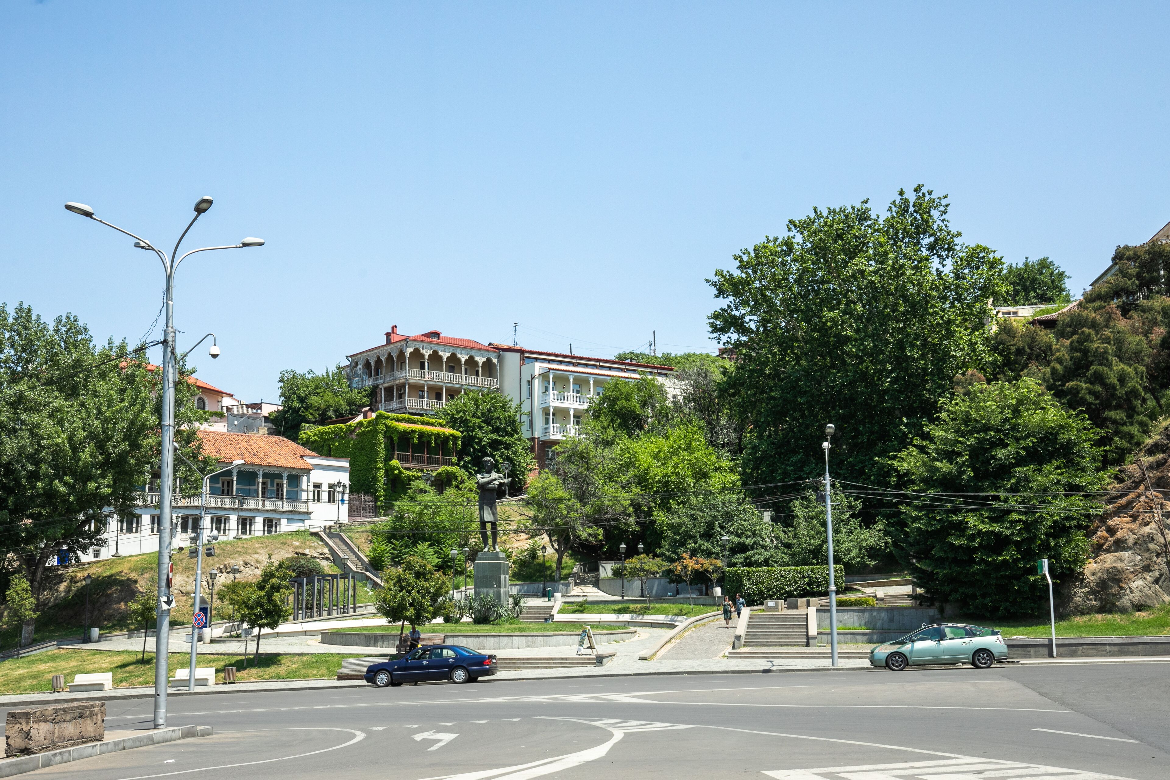 Vista de una calle con un edificio de techo rojo y un balcón, una estatua en el centro, y coches aparcados al borde de la carretera.