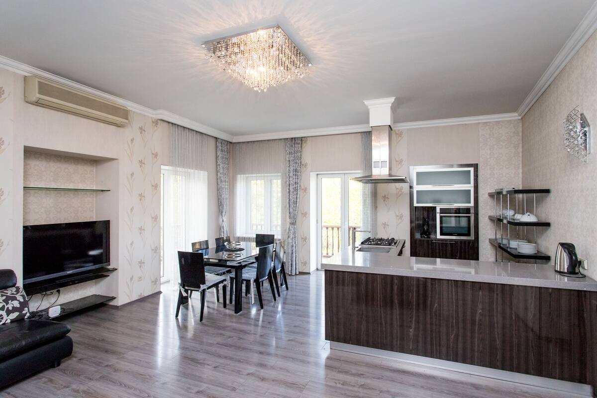 A modern living room with a flat-screen TV, a dining area with a table and chairs, a kitchen with a microwave and oven, and a chandelier.