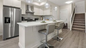 A modern kitchen with stainless steel appliances, white cabinetry, and a central island with bar stools. - 1BR Walk Out Suite » Backyard » Patio » 65” TV (Calgary)