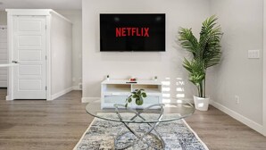 A modern living room with a glass coffee table, a white entertainment unit, a potted plant, and a wall-mounted TV displaying the Netflix logo. - 1BR Walk Out Suite » Backyard » Patio » 65” TV (Calgary)