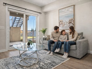 A living room with a glass coffee table, a grey sofa, and a large painting on the wall.