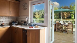 A kitchen with wooden cabinets, a sink, and a window overlooking a lakeside patio with outdoor seating. - Lake House -  Luxury Cottages (Brecon)