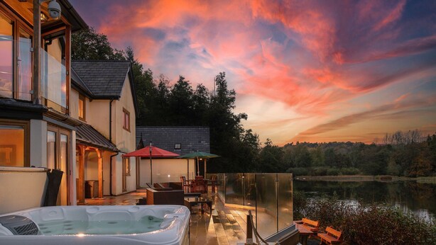 A hot tub with a glass enclosure, outdoor seating, and a view of a lake at sunset. - Lake House -  Luxury Cottages (Brecon)