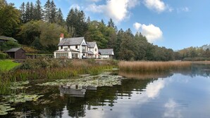 A lakeside house with a large deck, surrounded by dense forest and a calm lake. - Lake House -  Luxury Cottages (Brecon)
