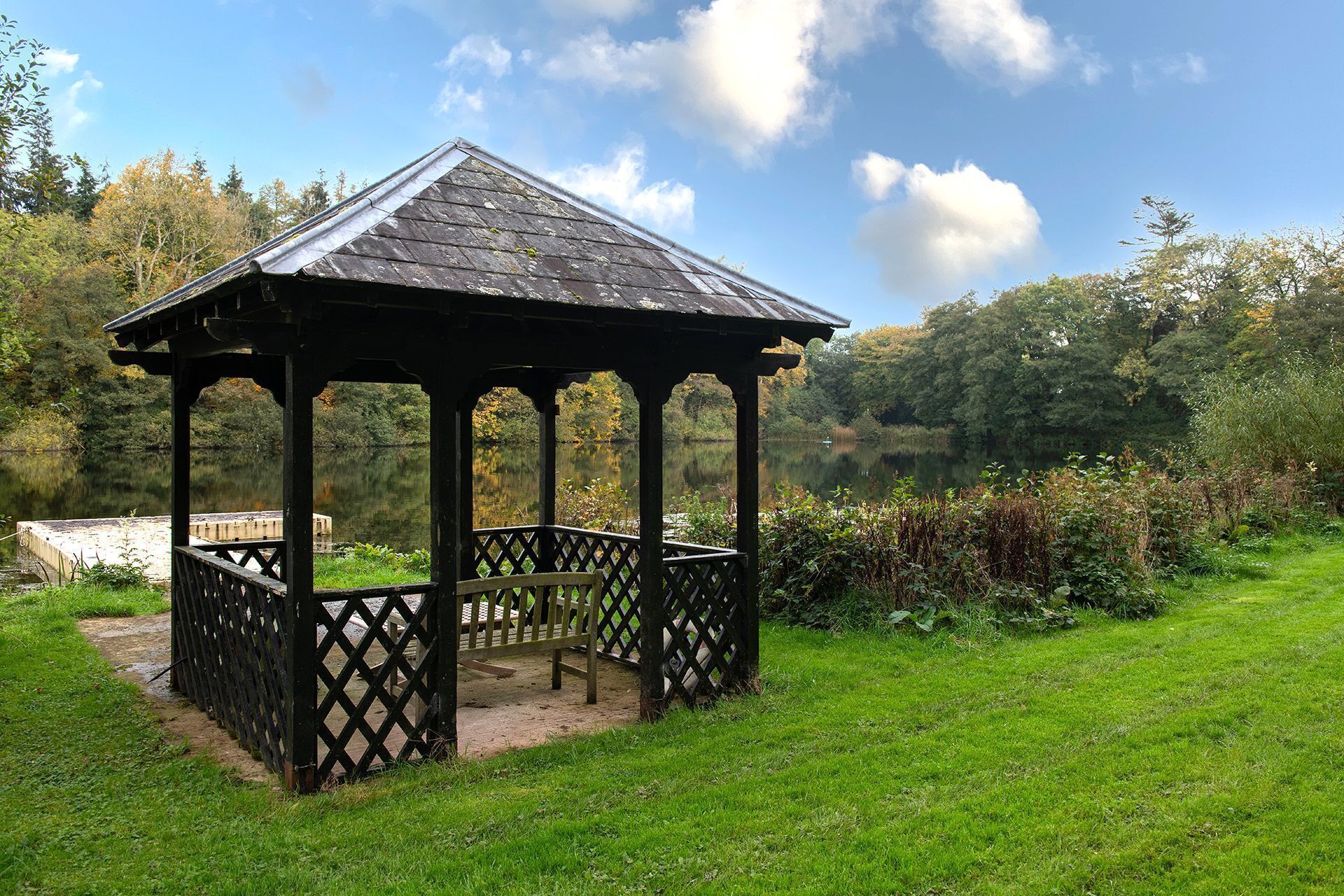 A gazebo with a wooden roof and lattice railings, situated on a grassy area near a body of water, surrounded by trees and vegetation.