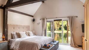A bedroom with a large bed, a bench with breakfast items, a view of a lake, and a wooden wardrobe. - Lake House -  Luxury Cottages (Brecon)