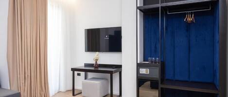 A modern hotel room with a flat-screen TV, a desk with a vase, a blue wardrobe, and a grey sofa.