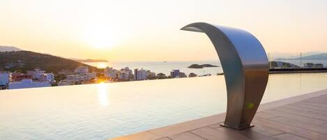A modern poolside area with a curved metal structure, overlooking a coastal cityscape at sunset.
