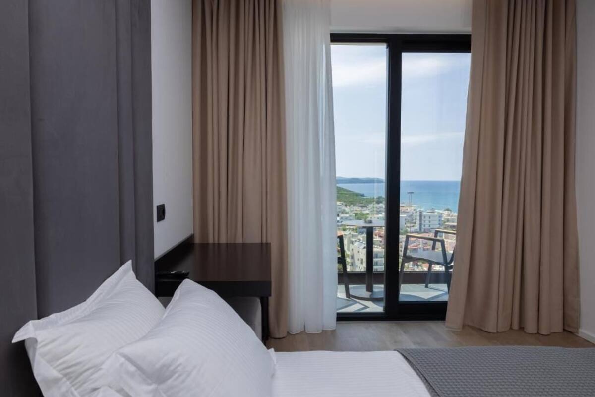 A bedroom with a bed, a desk, and a view of the sea and cityscape through a large window.