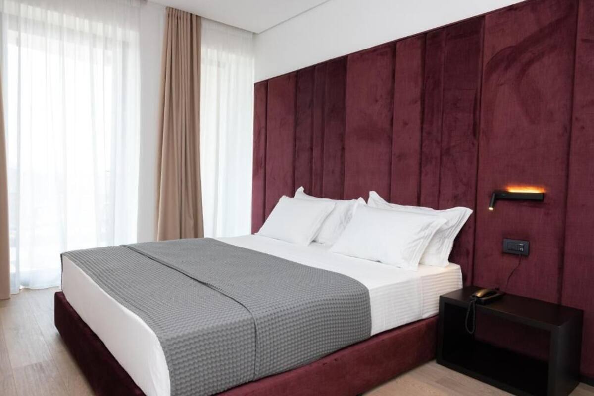 A neatly made bed with white linens and a grey blanket, a dark red headboard, and a bedside table with a phone.