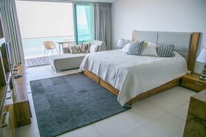 A modern hotel room with a large bed, a seating area with a view of the ocean, and a balcony with a table and chairs.