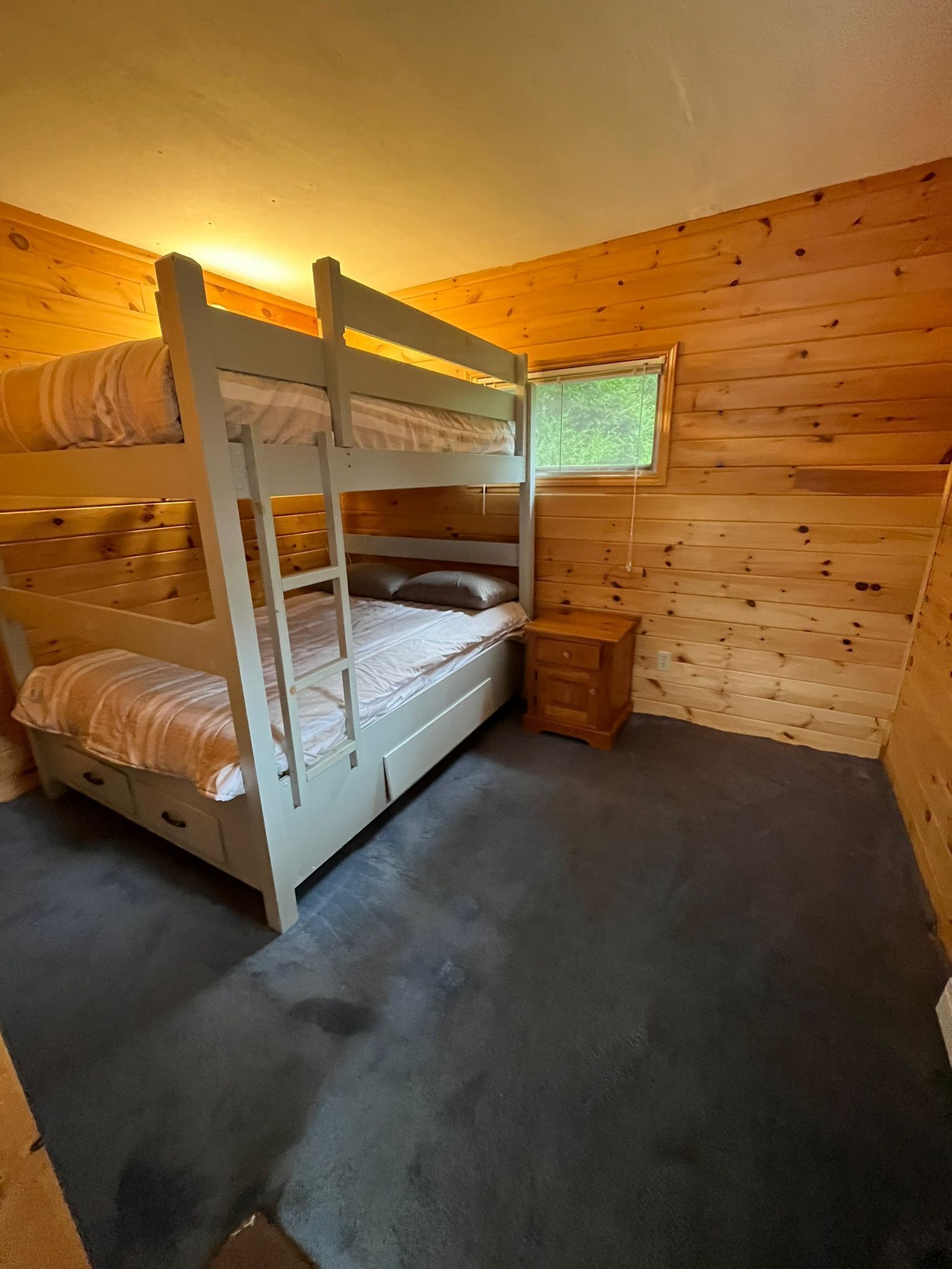 Ein Schlafraum mit Etagenbetten, einem kleinen Nachttisch aus Holz und einem Fenster mit Blick ins Grüne.