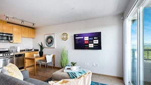 Una sala de estar moderna con un sofá gris, un comedor con una mesa y sillas de madera, una cocina con electrodomésticos de acero inoxidable y un televisor grande montado en la pared.