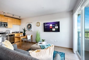 Un soggiorno moderno con un divano grigio, un'area pranzo con tavolo e sedie in legno, una cucina con elettrodomestici in acciaio inossidabile e un grande televisore a parete.