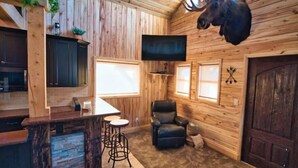 Een houten blokhut van binnen, met een bar, een opgehangen elandshoofd, een televisie en een gezellige zithoek.