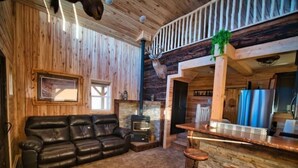 Een knus cabin interieur met een leren bank, een open haard, een bar met krukken en een houten plafond.