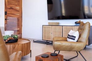 Una sala de estar moderna con un sillón de cuero, una mesita de madera, una mesa de centro redonda, un televisor de pantalla plana montado en la pared y un aparador de ratán.