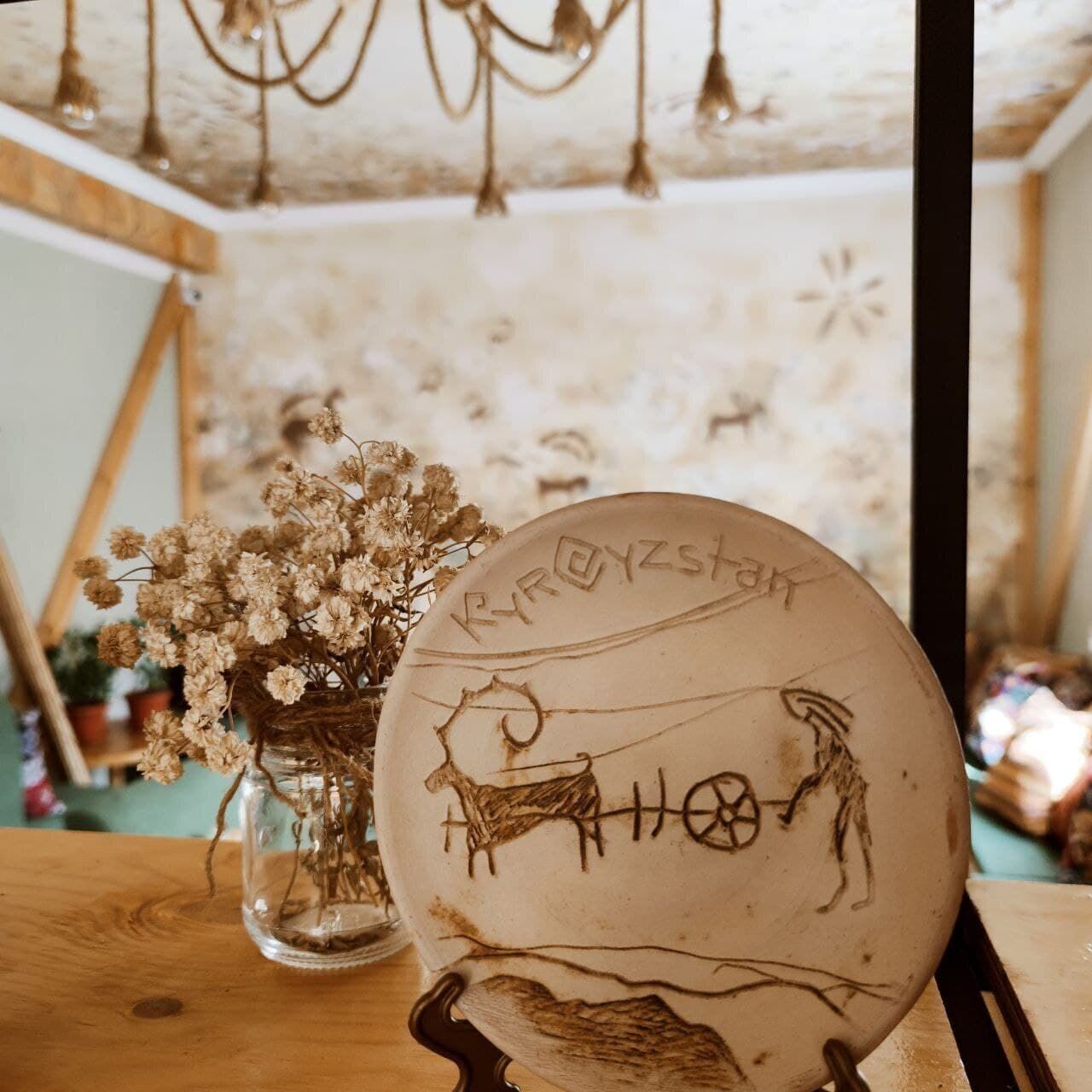 A rustic interior with a wooden table, a glass vase with dried flowers, and a ceramic plate with etched designs.