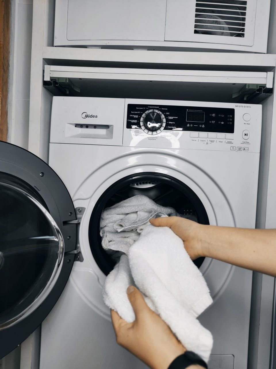 Une machine à laver à chargement frontal, dotée d’un panneau de commande, et une personne qui y place une serviette.