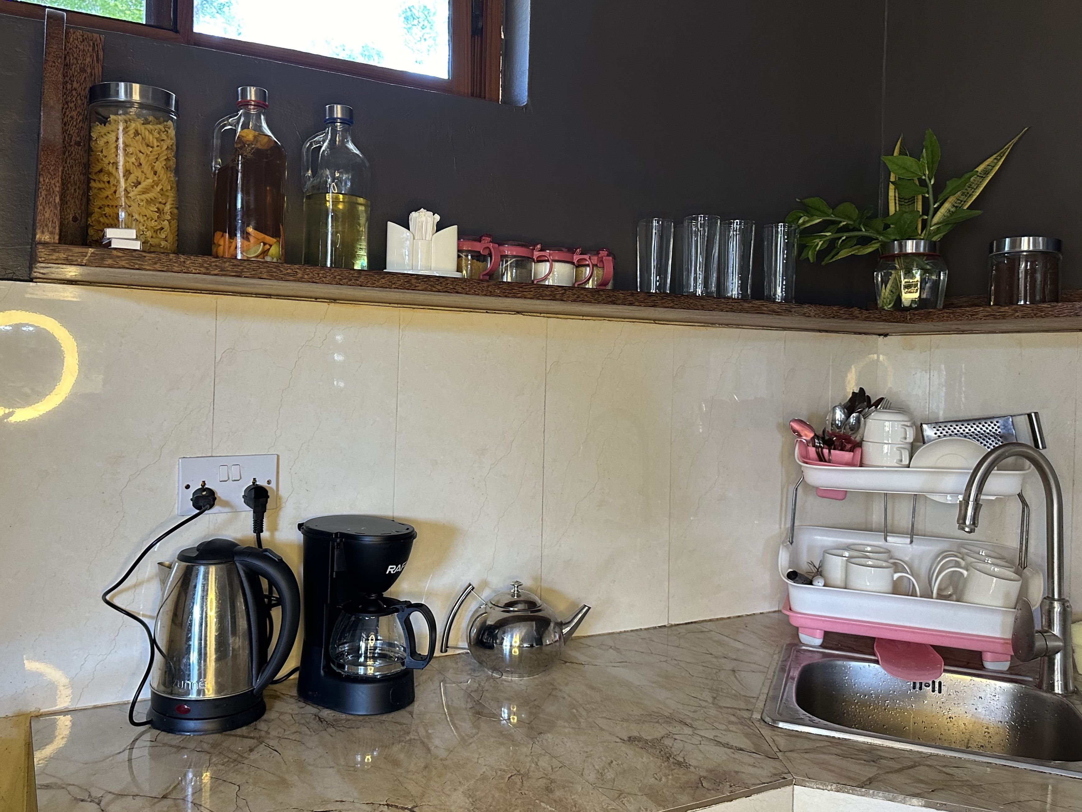 Een keuken met een marmeren aanrecht, een koffiezetapparaat, een waterkoker, een spoelbak met een afwassysteem, en diverse keukengerei en containers op de plank.