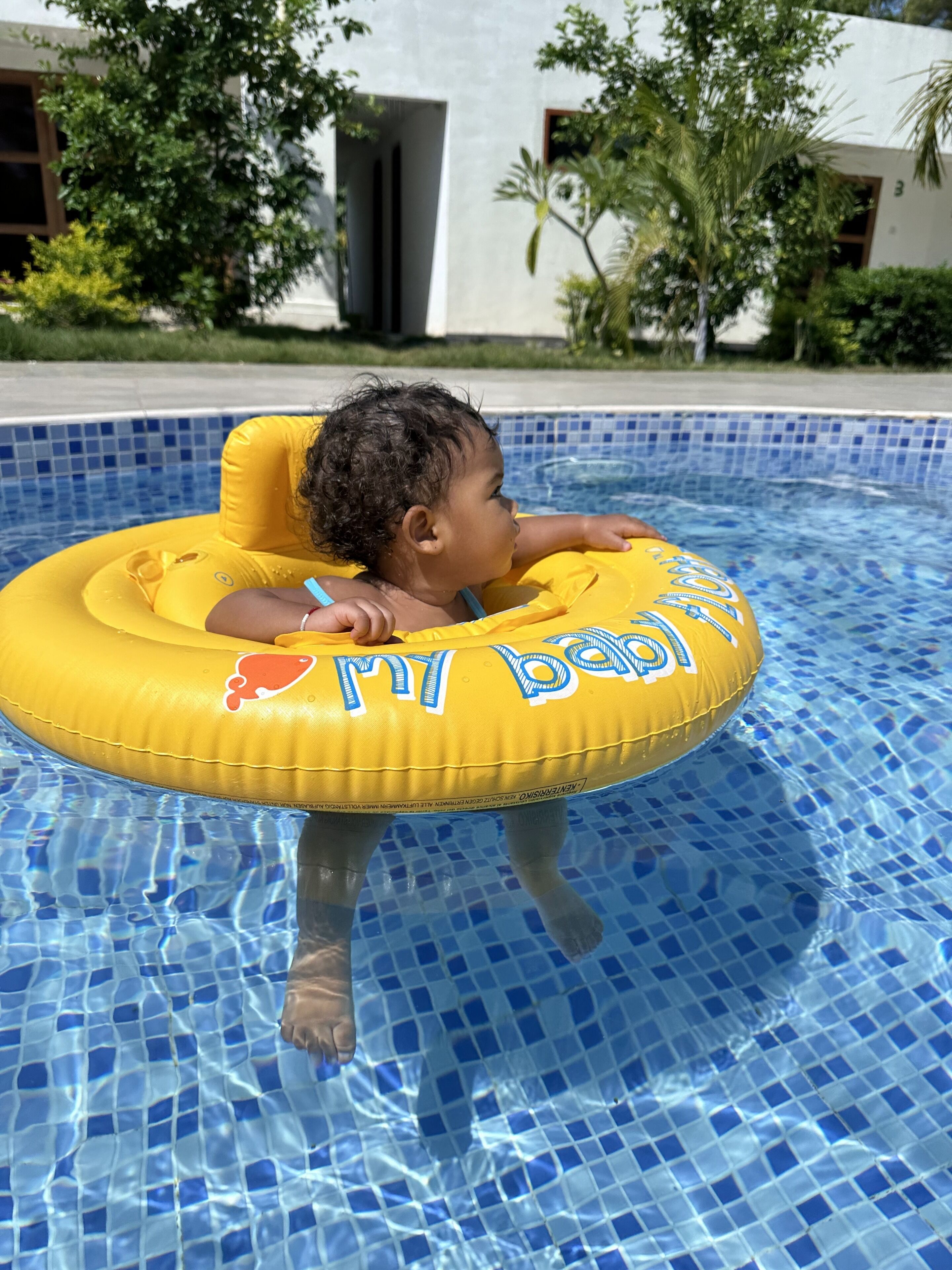 Un enfant dans une piscine gonflable jaune sur laquelle on peut lire les mots « My Baby », dans une piscine carrelée d’un bleu clair.