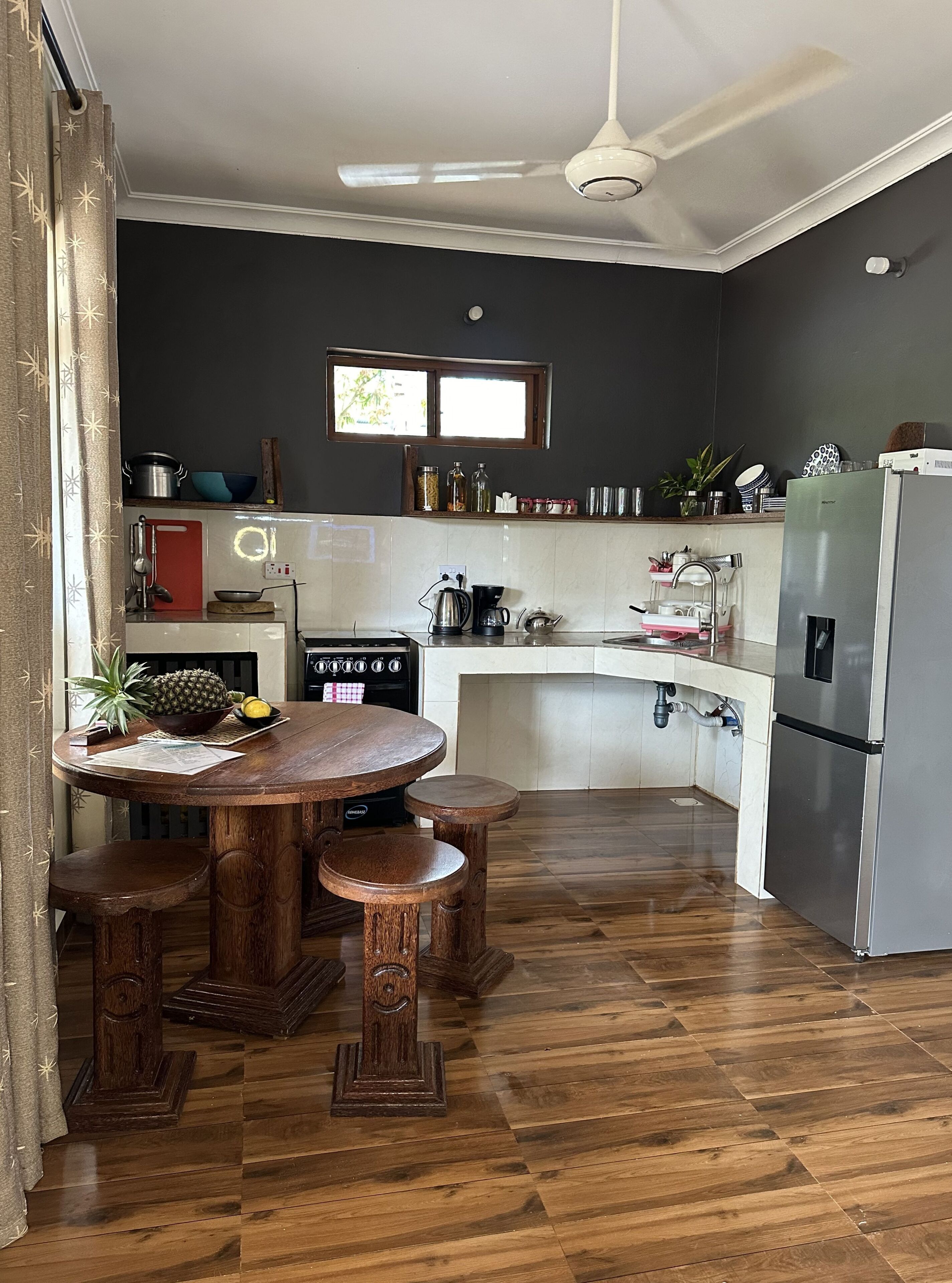 A modern kitchen with a wooden dining table, stools, a built-in oven, and a refrigerator.