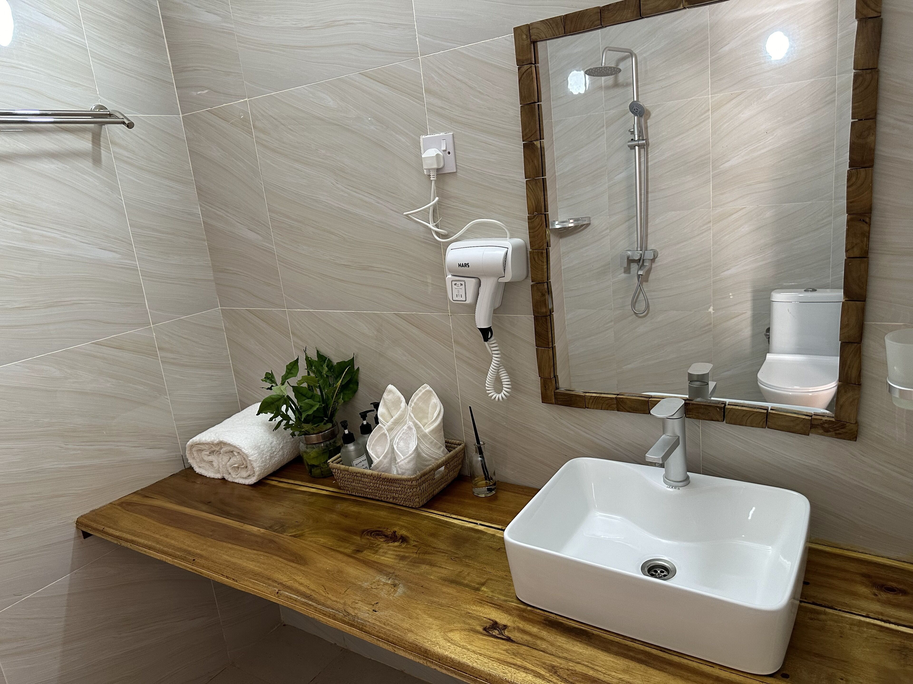 Un baño con tocador de madera, un lavamanos blanco, una toalla, una planta y un espejo con marco de madera.