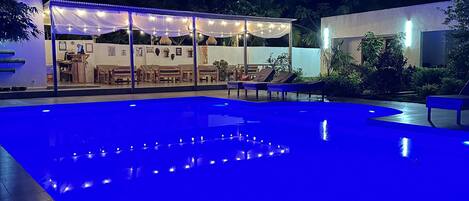 A swimming pool with blue lighting, a covered seating area with tables and chairs, and a modern building with a glass facade.
