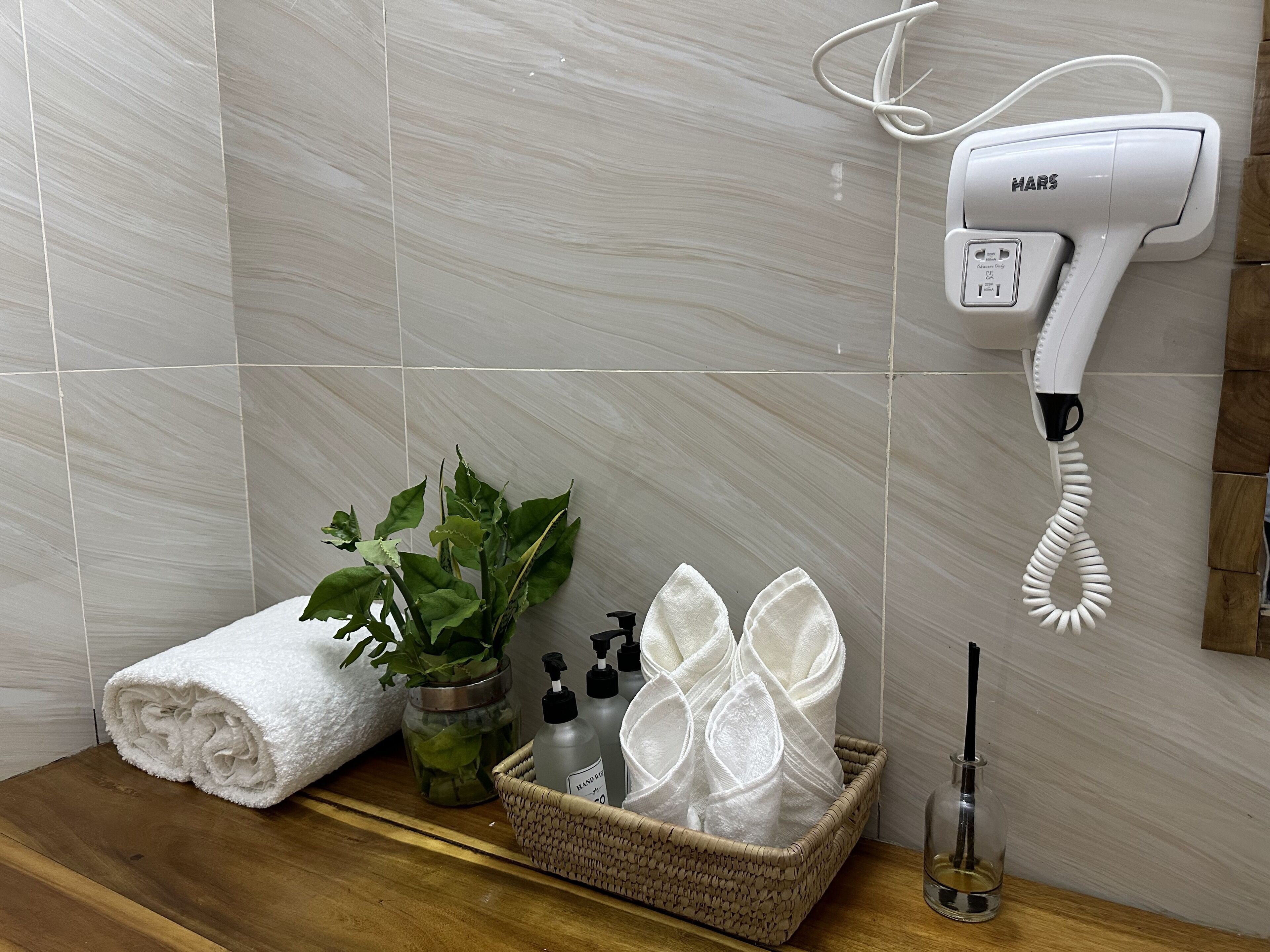 A bathroom with a hairdryer mounted on the wall, a rolled towel, a plant, a basket with folded towels, and a bottle of hand sanitizer.