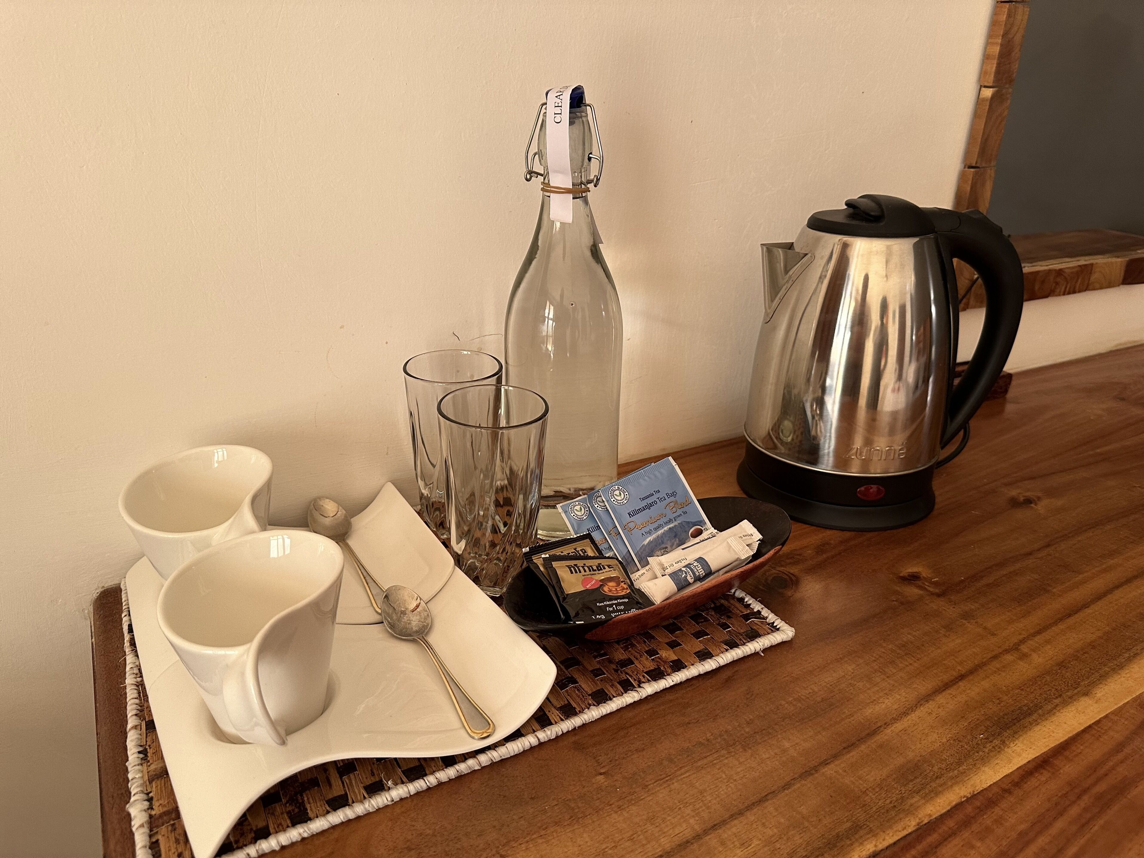 A wooden table with a clear bottle, two glasses, a white mug, a spoon, a packet of coffee, and a stainless steel kettle.