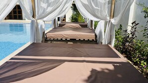 Eine Sonnenliege am Pool mit weißen Vorhängen, klares Wasser und viel Grün.