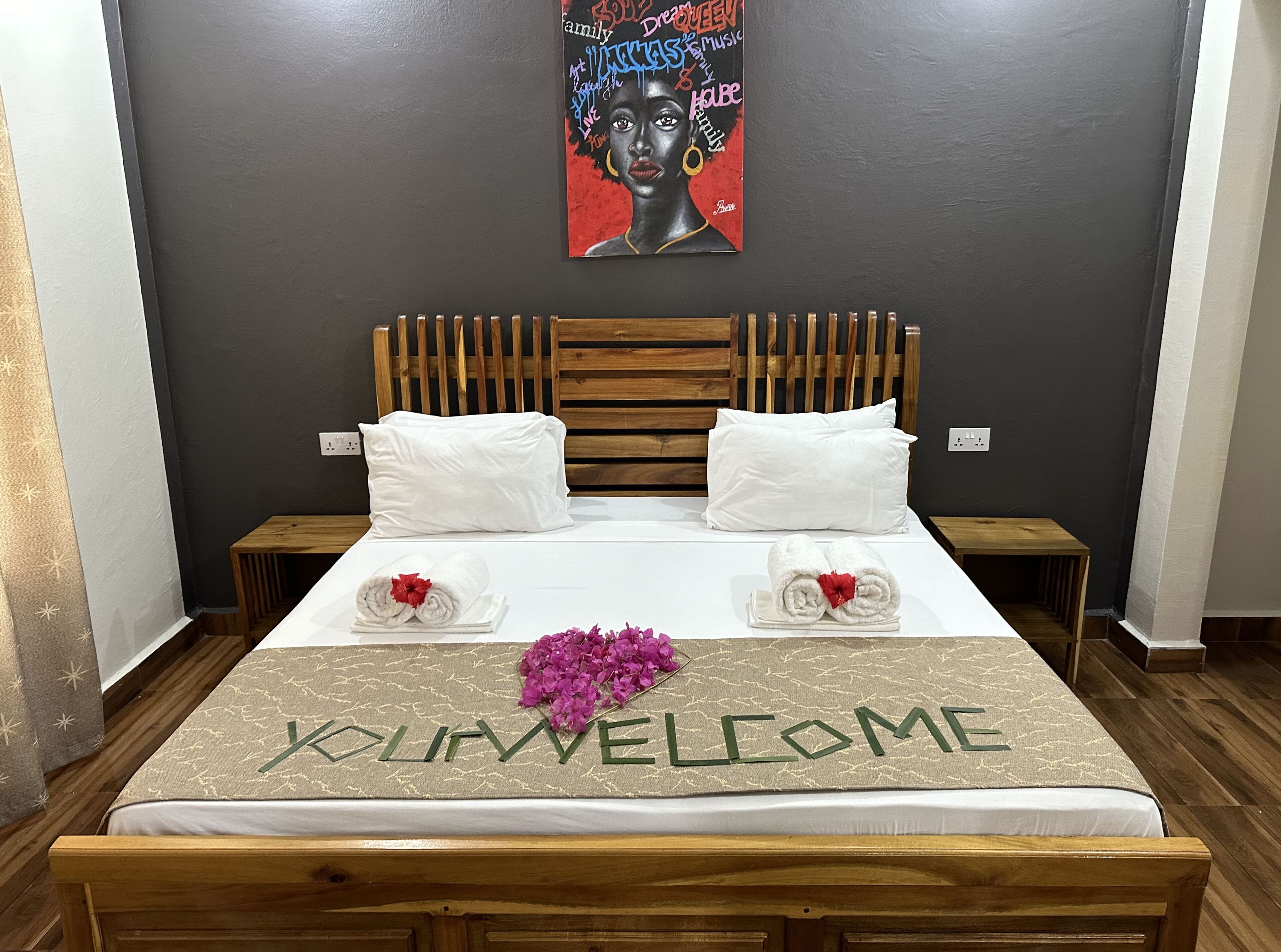 Una cama bien hecha, un tapete de bienvenida, dos almohadas y un arreglo floral decorativo.