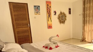 Ein ordentlich bezogenes Bett mit Kissen, eine dekorative Schwane Skulptur und Wandkunst, darunter ein Gemälde einer Frau auf einem Fahrrad und ein Spiegel in Sonnenform.