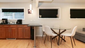 A modern kitchen with wooden cabinets, a dining area with a glass table and white chairs, and a grey sofa.