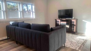 A living room with a dark-colored sofa, a flat-screen TV on a wooden stand, and a patterned rug on the wooden floor. - Silver Spring Tranquil Bliss-2BR/1BA Nature/Access (Silver Spring)