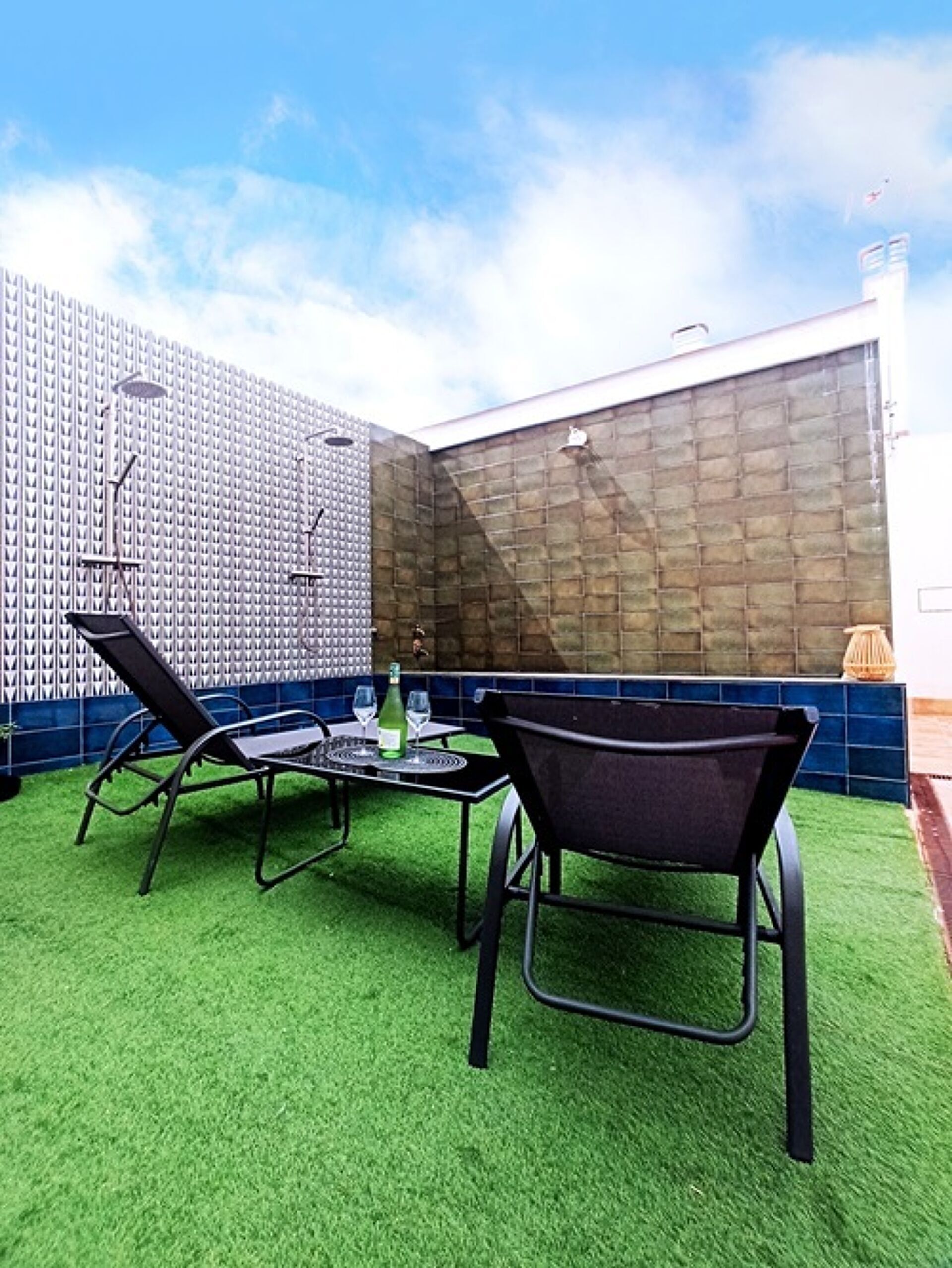 An outdoor patio with artificial grass, two black lounge chairs, a round table with a bottle and glasses, and a patterned wall.