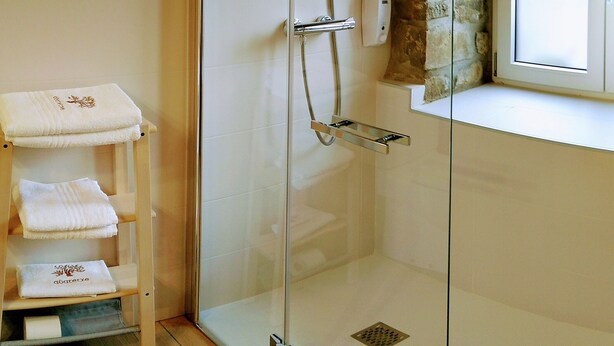 A glass-enclosed shower with a handheld showerhead, a towel rack with folded towels, and a stone wall adjacent to the shower. - Casa Rural Abatetxe (Elgoibar)
