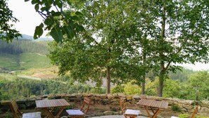 A seating area with four wooden chairs and tables, surrounded by a stone wall, overlooking a scenic valley with a road and forested hills. - Casa Rural Abatetxe (Elgoibar)