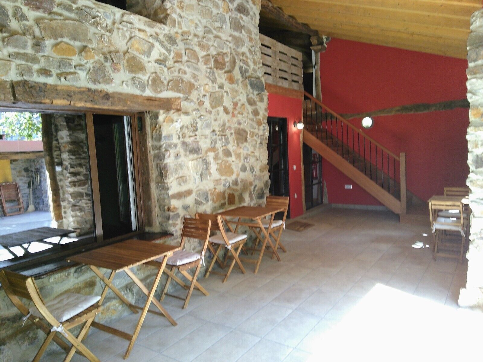 Una habitación con paredes de piedra, una mesa y sillas de madera, una escalera y una pared roja.