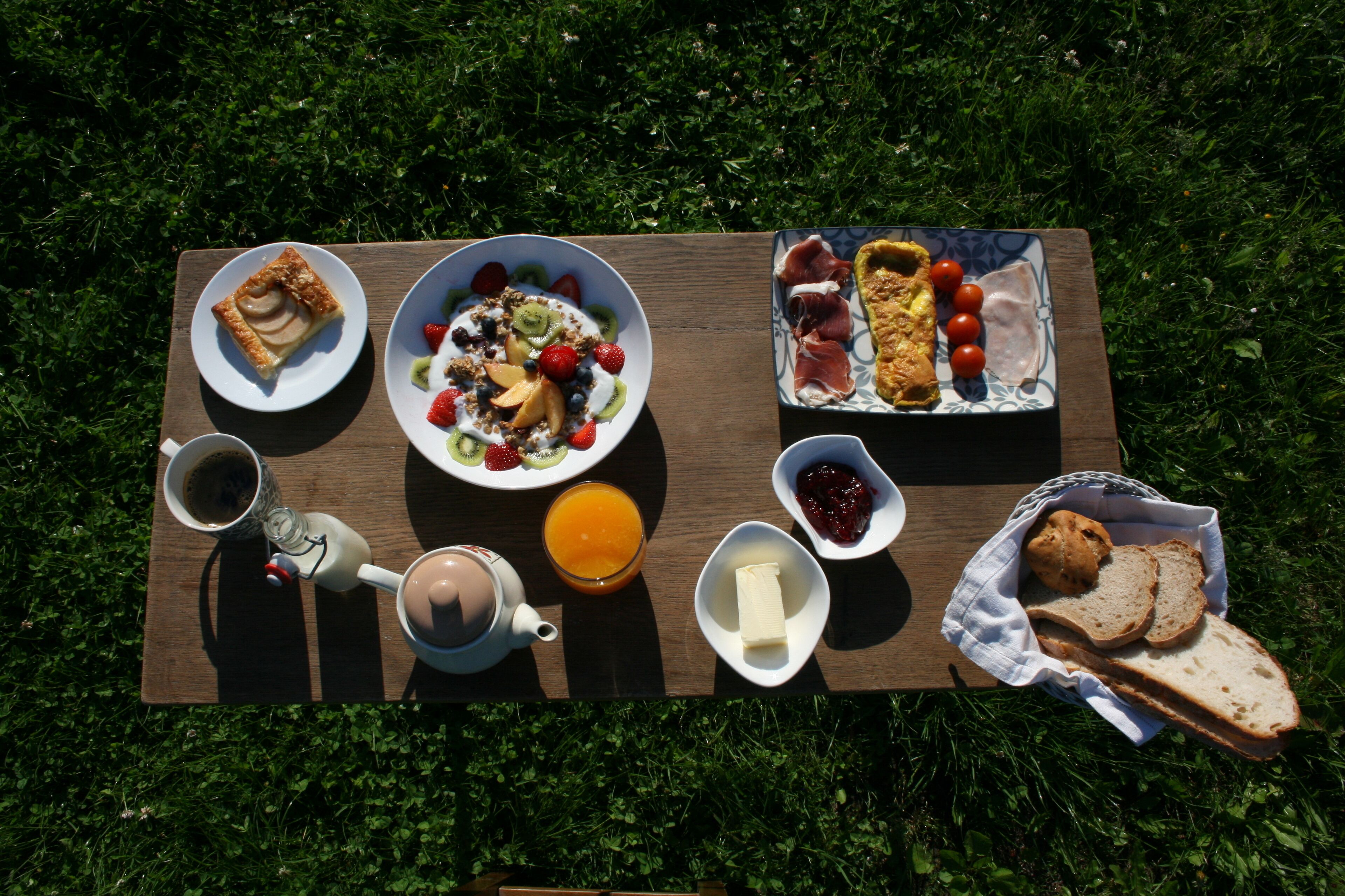A picnic setup on a grassy area with a variety of foods including a pastry, fruits, bread, and a beverage.