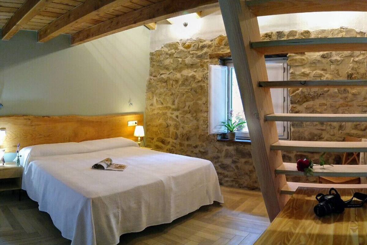 A bedroom with a bed, wooden headboard, stone wall, window, and a wooden table with a camera.