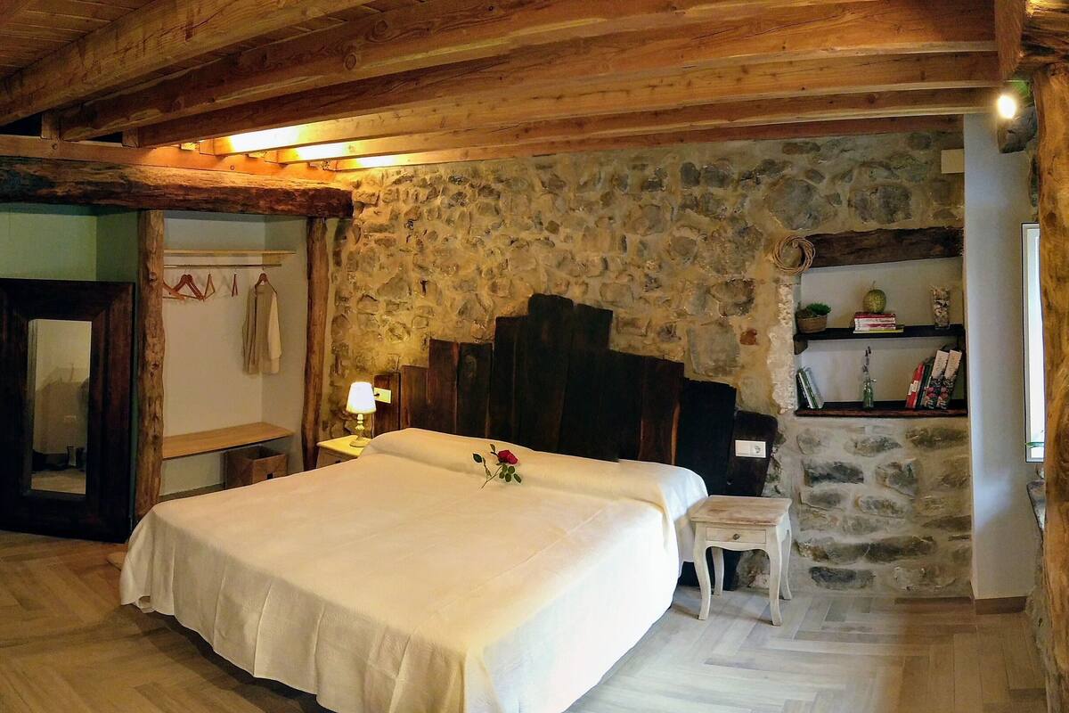A bedroom with a stone wall, wooden ceiling beams, a bed with a white bedspread, a bedside table with a lamp, and a small stool.