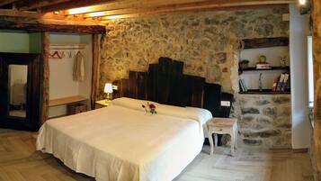 A bedroom with a stone wall, wooden ceiling beams, a bed with a white bedspread, a bedside table with a lamp, and a small stool.